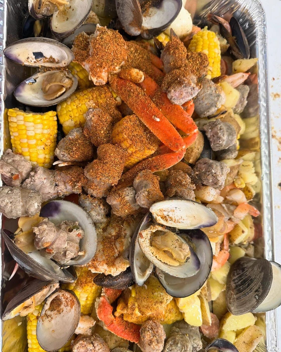 A seafood boil packed in a foil tray: snow crab, clams, shrimp, sausage, corn and potatoes under our own boil spice